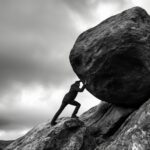 Silhouette man struggles to push massive boulder uphill on rocky terrain image. Overcast sky, rugged landscape photography scene wallpaper. Sisyphean task resilience concept photo